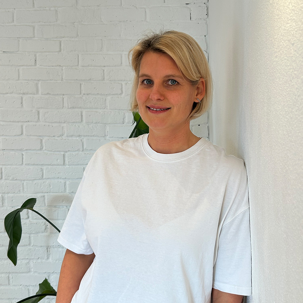 Eine lächelnde Frau mit blondem Haar, weißes T-Shirt, steht vor einer weißen Backsteinwand.