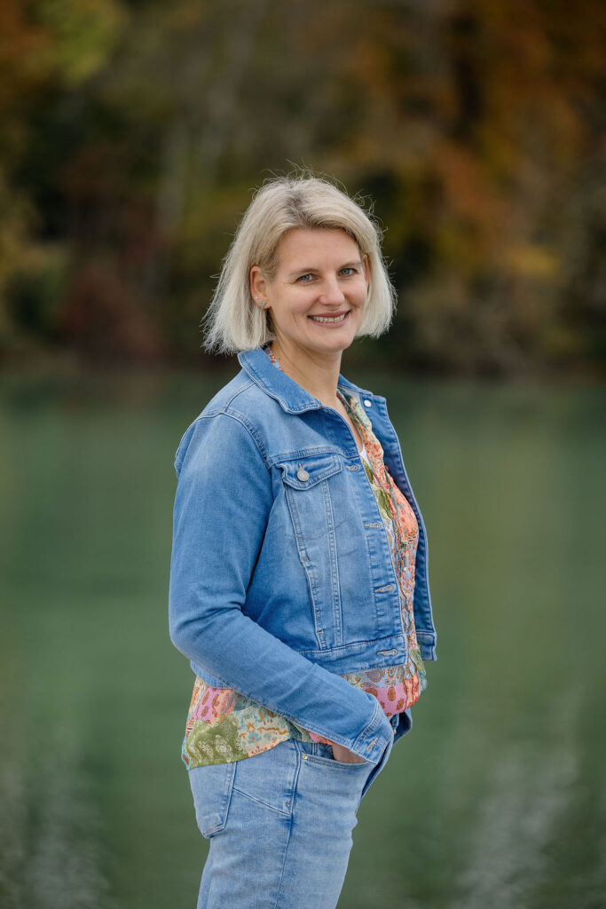 Eine lächelnde Frau mit blonden Haaren trägt eine blaue Jeansjacke vor grünem Wasser